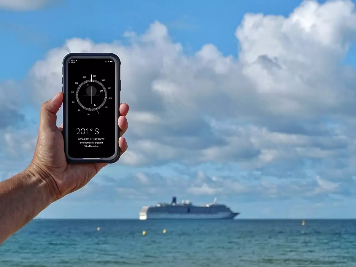 A hand holds an iPhone against a backdrop of a vast ocean, a large cruise ship on the horizon, and a blue sky with fluffy clouds.